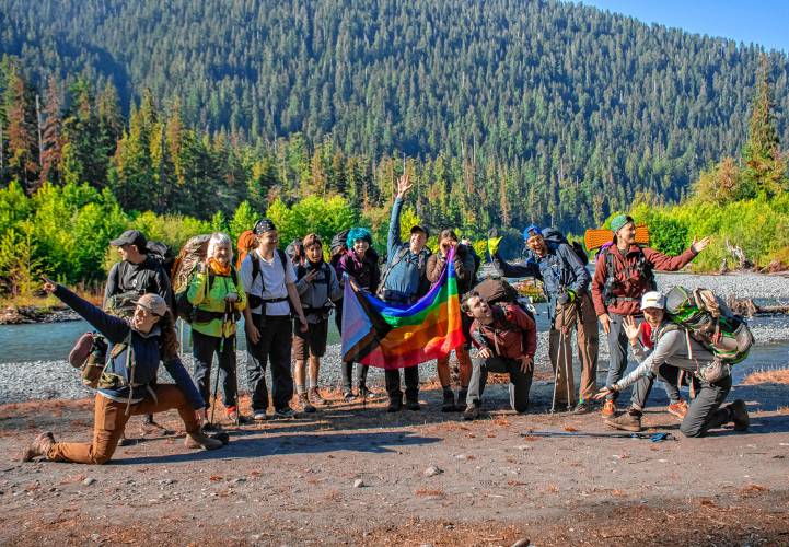 Twelve queer people wearing outdoor gear and backpacks pose together in the sun, holding a large progress pride flag in the center. They’re lined up in front of a rocky blue river beneath a conifer forest.