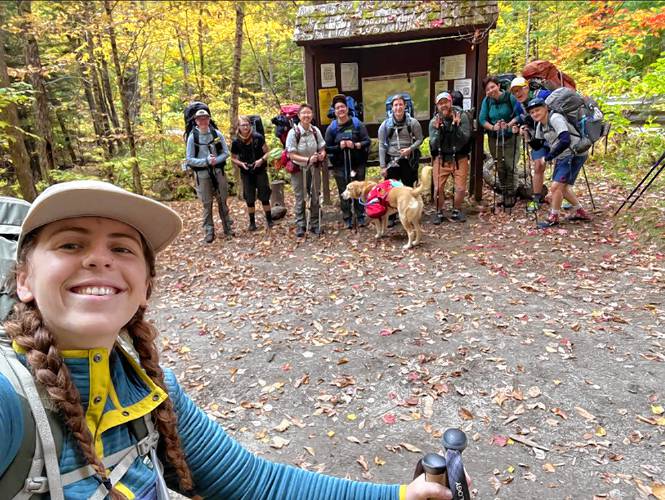 The Venture Out Project’s Ana Seiler smiles for a selfie with nine queer backpackers and one dog all dressed for a backcountry trip standing behind her in the autumn leaves.
